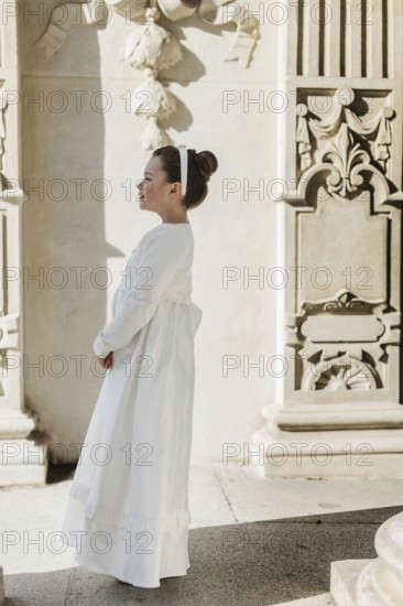 A young girl wearing a First Communion dress stands by an ornate wall. The scene captures a serene
