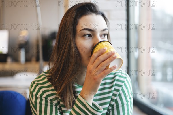 A young woman in a striped shirt takes a peaceful coffee break, sipping from a yellow cup inside a