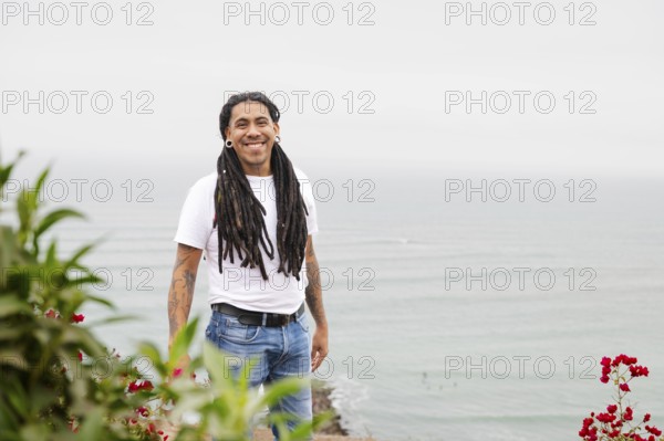 A man with long dreadlocks and tattoos smiles warmly, standing near vibrant flowers with the serene
