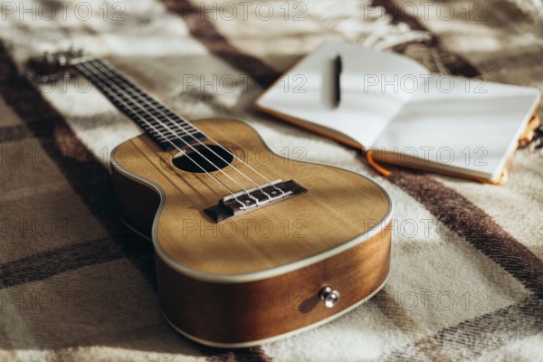 A ukulele rests on a cozy, plaid blanket beside an open notebook with a pen, capturing a serene,