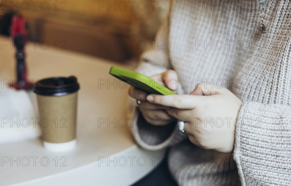 Close-up of a woman's hands holding a green smartphone in a bookstore cafe A takeaway coffee cup