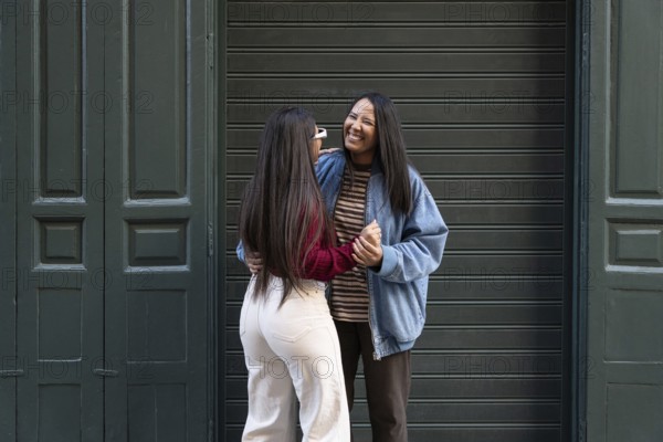 A joyful Latin lesbian couple stands in an embrace, laughing together in front of a green door.