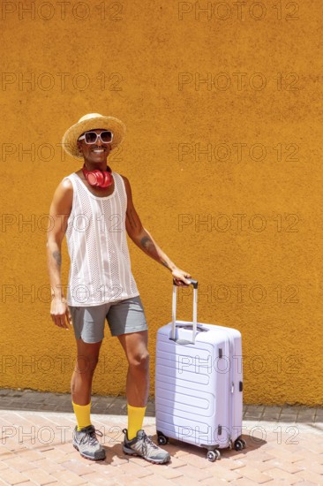LGBTQIA+ man stands joyfully beside a suitcase, dressed vibrantly and ready for a holiday. A