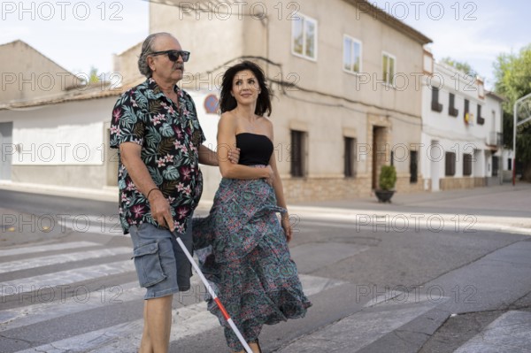 A blind father uses a white cane while his daughter guides him through a tranquil town street. Both