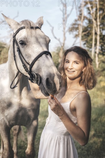 Young woman in a white dress gently pets a white horse on a sunny day. The serene outdoor setting
