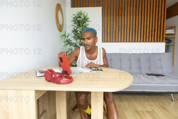 A Hispanic LGBTQIA+ man with vibrant style sits at a table, utilizing a smartphone and maps for