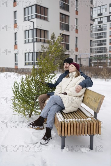 A couple sits on a wooden bench surrounded by snow, holding a small Christmas fir They are bundled