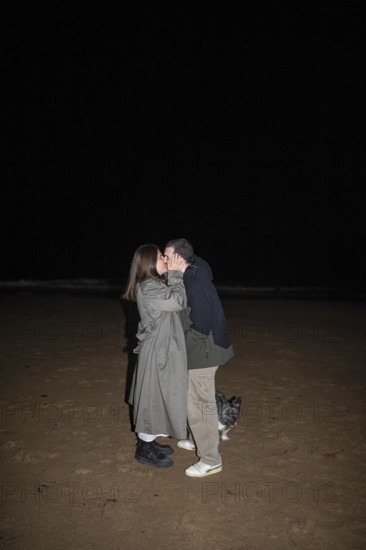 A couple shares a tender kiss under the night sky on a sandy beach, accompanied by a small dog The