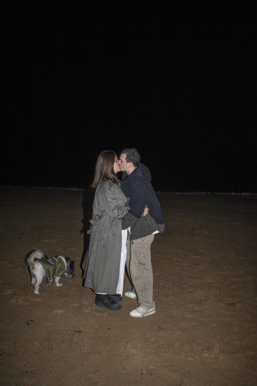 A couple shares a tender kiss on a dark beach night, their small dog beside them The scene captures