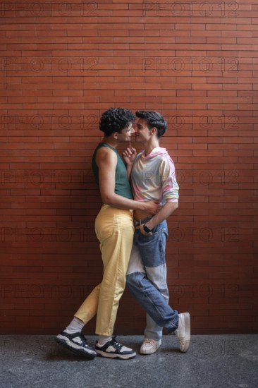 A young gay couple shares an intimate moment against a brick wall, expressing affection and love.