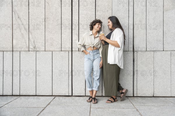 A relaxed lesbian couple enjoys a casual moment together, leaning against a modern stone wall. Both
