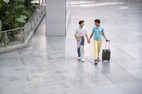 A joyful gay couple walks hand in hand through an airport terminal with luggage, enjoying each