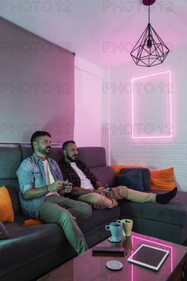 A gay couple relaxes on a sofa, playing video games in a warmly lit living space with modern decor.