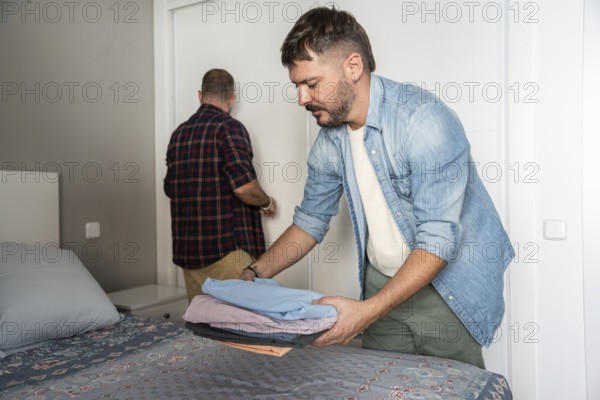 A gay couple organizes clothes and tidies up their bedroom. One is folding laundry on the bed while