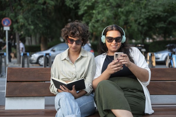 A lesbian couple enjoys a sunny day outdoors, sitting on a bench. One is reading a book, and the