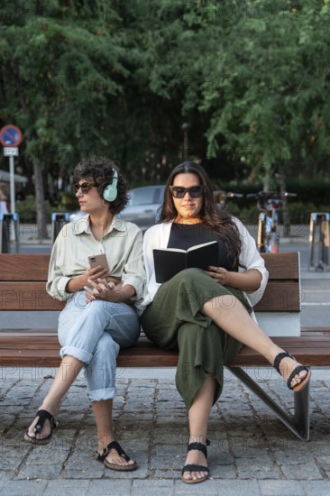 Lesbian couple relaxes on a park bench. One is reading a book, while the other listens to music,