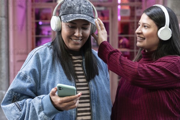 A smiling Latin lesbian couple enjoying music together with headphones. One holds a smartphone,