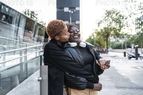 A loving LGBT multiethnic couple is embracing and smiling on an urban street. One partner gently
