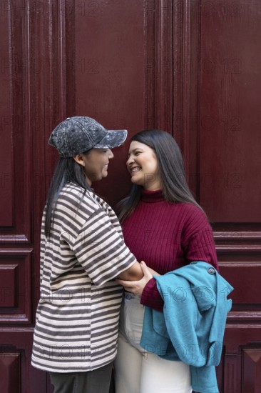 A joyful Latin lesbian couple in a loving embrace stands against a deep burgundy wooden backdrop,