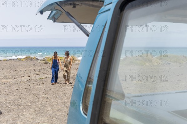 A lesbian couple strolls hand in hand along a beach, adjacent to a classic van. They enjoy the