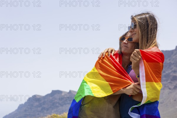Two women embrace under a colorful rainbow flag, symbolizing LGBTQIA pride and unity, set against a