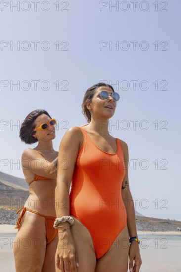 Lesbian women in stylish orange swimsuits enjoying a sunny beach day. They wear sunglasses, showing