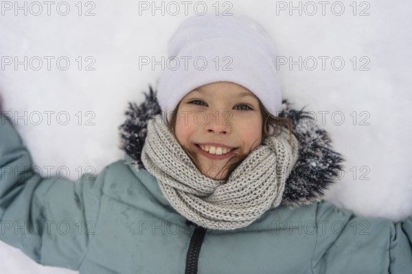 A smiling child lies in the snow, wearing a warm jacket, white hat, and knitted scarf. The joy and