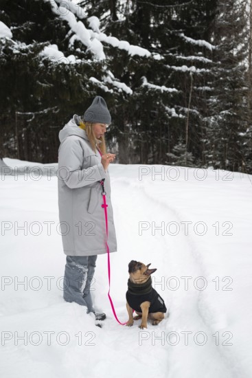A woman in a warm coat and hat walks her dog on a snowy trail. The forest around is covered in