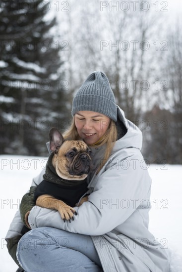 A woman in winter attire lovingly holds her French Bulldog in a snowy landscape. The scene captures