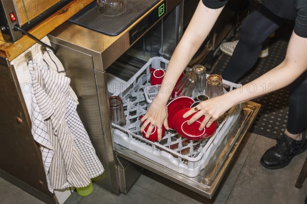 A person loads a dishwasher with red coffee cups and glass bottles, capturing the behind-the-scenes