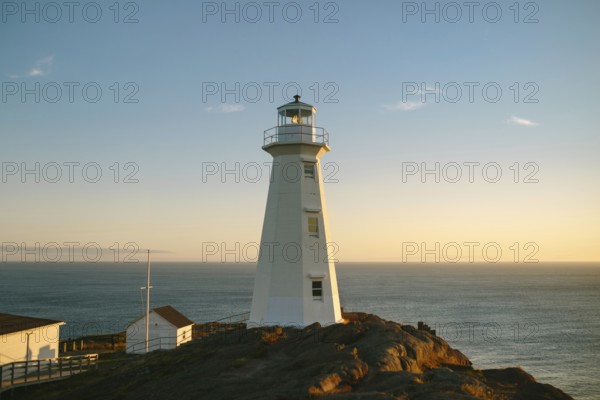 A warm sunset casts a golden glow over the white structure of Cape Spear Lighthouse, perceived as
