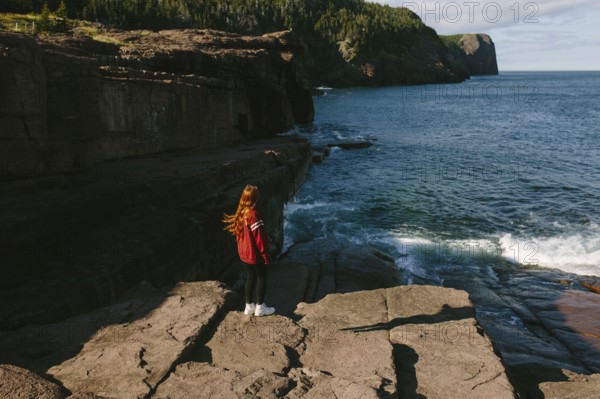 Back view of a young unrecognizable redhead woman standing on the rocky shoreline of Flatrock,