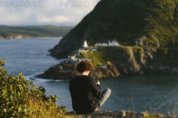 Back view of an unrecognizable man sitting atop a rocky cliff at St. John's, Newfoundland &
