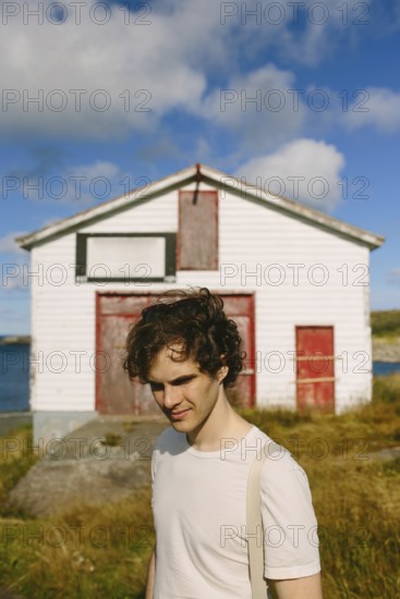 A young man with curly hair stands outdoors, smiling subtly near an old white building with red