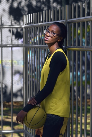 A woman basketball player in a yellow jersey and glasses leans against a metal fence, holding a