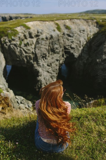 Back view of an unrecognizable redhead woman sitting on a grassy edge, overlooking a rugged coastal