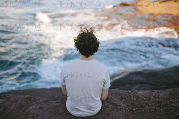 Back view of an unrecognizable person sitting on a rock by the sea, enjoying the serene view at