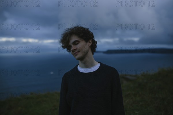 A thoughtful young man with eyes closed stands by the ocean in Signal Hill in St. John's,
