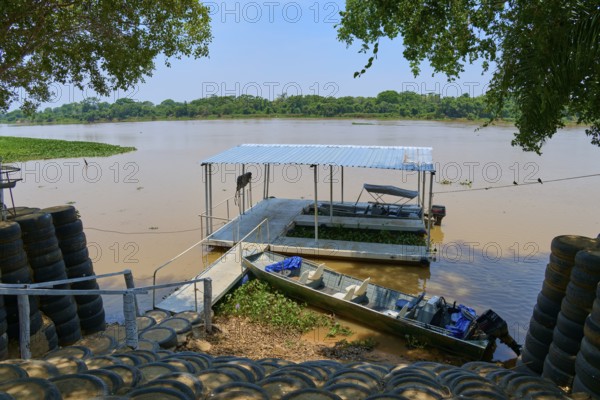 A jetty stretches across the river with a small boat nearby, surrounded by trees, Porto Jofre,