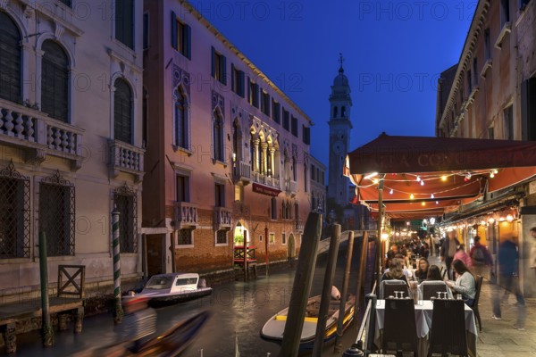 In the evening, lively restaurant on a canal with the tower of Santa Maria della Pietà in the back,