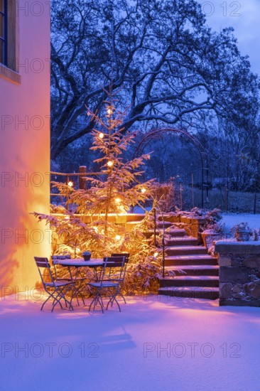 Still life with illuminated Christmas tree, stairs and table with chairs in the snow at blue hour,