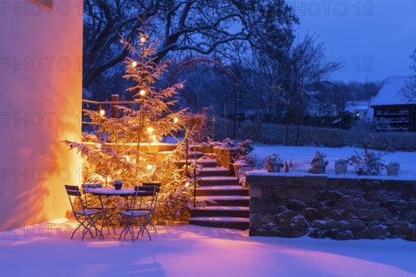 Still life with illuminated Christmas tree, stairs and table with chairs in the snow at blue hour,