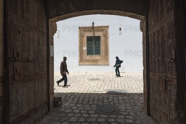 Tunisian men and women walk past some old wooden doors beside Bab Diwan the gate to Sfax Medina,