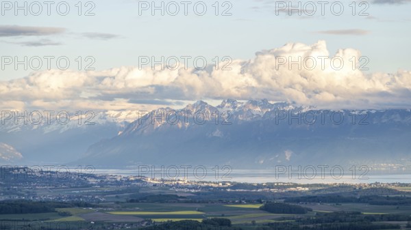 Lake Geneva stretches along the base of towering alpine mountains, with fluffy white clouds casting