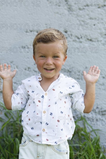A cheerful child with an adorable smile raises their hands playfully against a textured wall