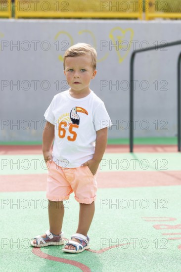 A young boy stands confidently on a vibrant playground, wearing a white T-shirt and peach shorts
