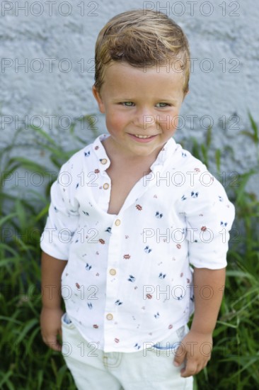 A cheerful young boy with a charming smile stands against a gray wall, dressed in a playful