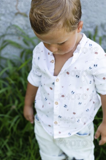 A young boy wearing a white shirt with colorful prints stands amidst green foliage His gaze is