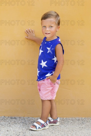 A young child stands against a vibrant orange wall, wearing a blue sleeveless shirt with white
