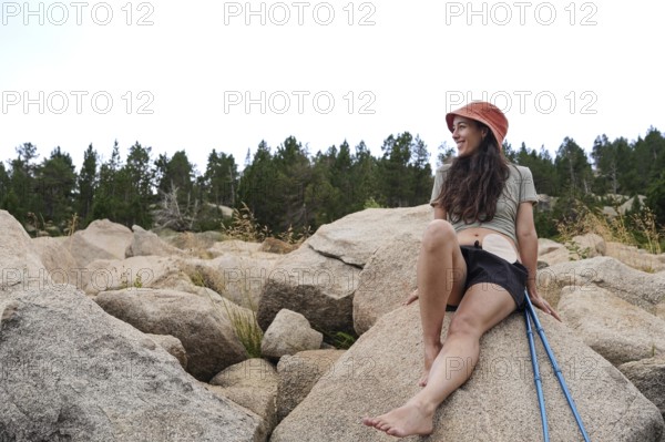 A smiling woman with an ostomy enjoys nature, sitting on large rocks amidst a serene forest